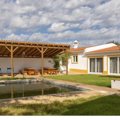 A modern villa with a pool, wooden terrace, and lush green garden under a cloudy sky.